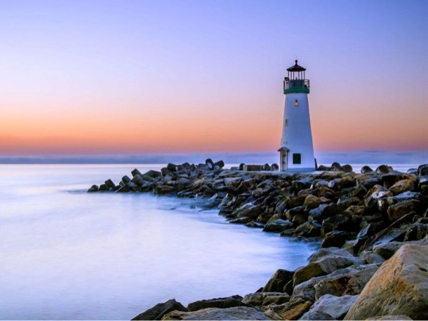 A lighthouse stands on the rocks, illuminated by the golden light of sunset.