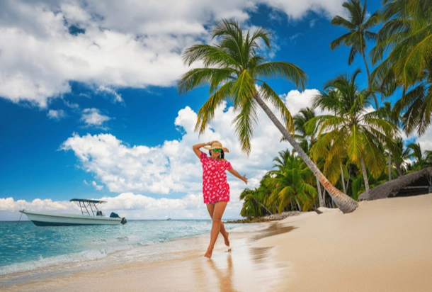 The image shows a person standing on a sandy beach looking out at the sea. There are palm trees, a clear blue sky, and a boat in the distance. This image depicts a serene and picturesque tropical beach scene, which might be relevant for topics related to travel, holidays, or nature. user imagen describi ingles dos lineas