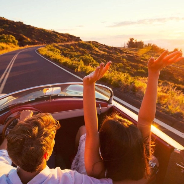 A smiling couple drive a convertible, hands raised, enjoying a sunny day.