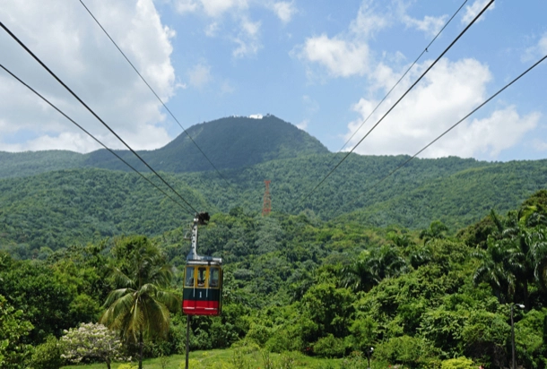 The image shows a vibrant green mountain landscape with a clear blue sky. In the foreground, there is a red and green cable car suspended on wires, which adds an element of human transportation and tourism interest to the natural scene. user imagen describi ingles dos lineas