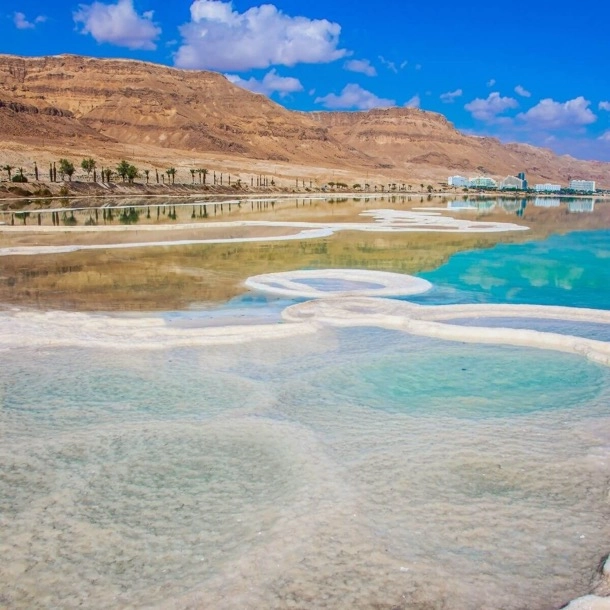 The image shows the Dead Sea with its characteristic salt formations and crystal-clear blue waters against a backdrop of arid hills under a blue sky. This scene is interesting due to the unique geological features of the Dead Sea, known for its high salinity and mineral-rich waters. 