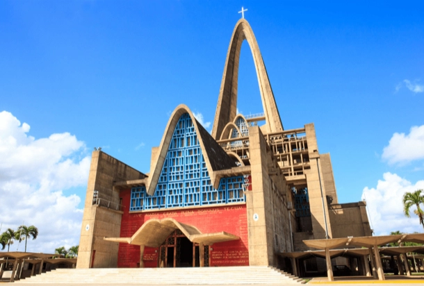 The image shows a modern architectural structure that appears to be a church, with distinctive pointed arches and a large cross at the top. The building has a unique combination of concrete and blue glass panels, set against a backdrop of a clear sky with few clouds. This image is interesting due to the contemporary design of the religious building, which stands out from traditional church architecture.