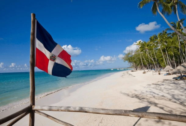 The image shows a beach with white sand and a clear blue sky. A flag, which appears to be the flag of the Dominican Republic, is prominently displayed in the foreground on a pole. The beach is lined with palm trees, suggesting a tropical location. This image might be interesting or relevant for discussions about travel, tropical destinations, or the Dominican Republic itself.