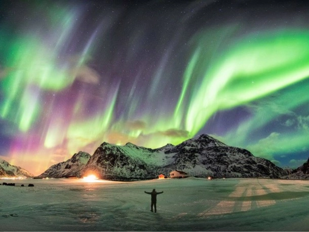 A person standing in the snow under a green and purple aurora borealis illuminating the night sky.